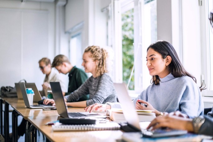 A group of students seated at a long table, working on laptops and notebooks. The classroom setting is bright, with large windows providing natural light, fostering a collaborative learning environment.